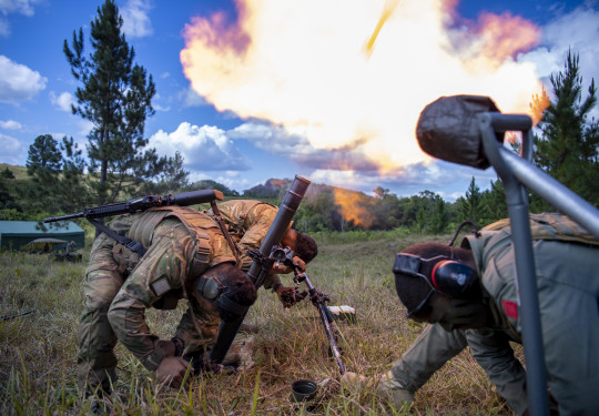 Three soldiers wearing hearing protection lower their heads as they fire a mortar, this creates a large flame . There are trees in the background and blue skies with scattered cloud.