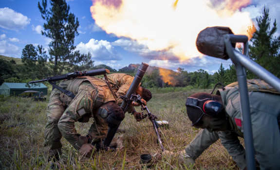 Three soldiers wearing hearing protection lower their heads as they fire a mortar, this creates a large flame . There are trees in the background and blue skies with scattered cloud.