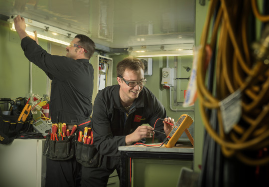 Two men wearing dark overalls, safety glasses and a tool belt work on electronics in an industrial room.