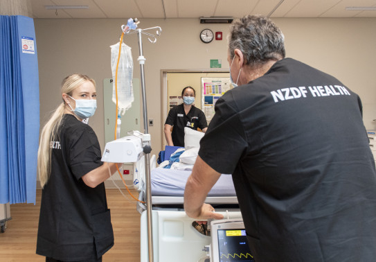 Three nurses move a medical bed in a hospital environment. They are all wearing black scrubs that read 'NZDF HEALTH' on the back. In the background is a doorway to a hall and there is a blue curtain hanging from the ceiling on the left.