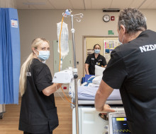 Three nurses move a medical bed in a hospital environment. They are all wearing black scrubs that read 'NZDF HEALTH' on the back. In the background is a doorway to a hall and there is a blue curtain hanging from the ceiling on the left.