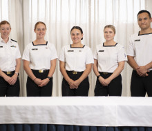 Five sailors wearing white tops and black trousers stand behind a table, in front of curtains.