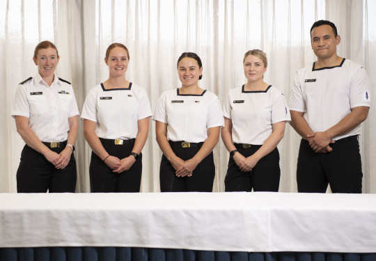 Five sailors wearing white tops and black trousers stand behind a table, in front of curtains.