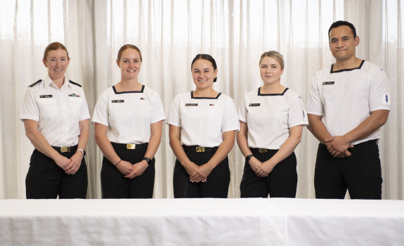 Five sailors wearing white tops and black trousers stand behind a table, in front of curtains.