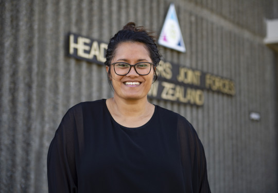 Miss Keri Brooking, 2022 Civilian of the Year, stands in front of the Headquarters Joint Forces New Zealand sign. She is wearing a black top and is smiling at the camera. 