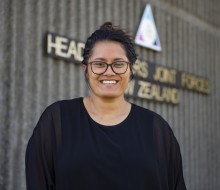 Miss Keri Brooking, 2022 Civilian of the Year, stands in front of the Headquarters Joint Forces New Zealand sign. She is wearing a black top and is smiling at the camera. 
