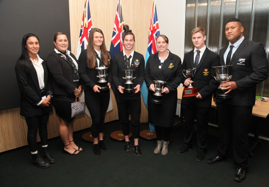 Seven award winners wearing black blazers, dresses and pants and white shirts stand in a line, they smile at the camera with their awards.