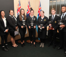 Seven award winners wearing black blazers, dresses and pants and white shirts stand in a line, they smile at the camera with their awards.