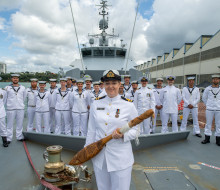 Woman in white navy uniform stands on the deck of HMNZS Taupo, a grey patrol ship with the crew, also in white dress uniform behind her.