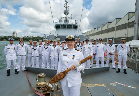 Woman in white navy uniform stands on the deck of HMNZS Taupo, a grey patrol ship with the crew, also in white dress uniform behind her.