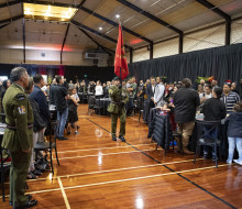 A soldier carrying a red flag walks down the centre of a hall as people stand next to the tables they were sitting at.