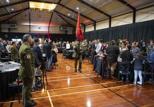 A soldier carrying a red flag walks down the centre of a hall as people stand next to the tables they were sitting at.