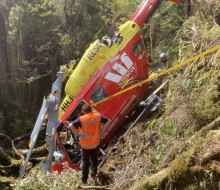 A red and yellow Westpac Rescue helicopter crashed in dense bush with a person inspecting the helicopter in the foreground. The area of bush around the helicopter has been cut and cleared, ready for recovery.