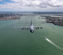 The grey C-130H Hercules aircraft flies over Auckland Harbour with the Auckland Harbour Bridge and SkyTower in the background.