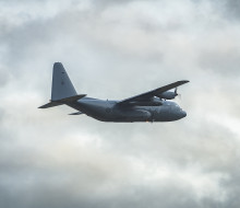 A rear-side view of the grey C-130 Hercules aircraft, with landing gear up shortly after taking off from Christchurch Airport. The clouds in the background are very textured and broken with a soft light glowing the edges.