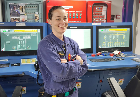 Able Marine Technician (Electrical) Gemma Townshend hold some tools in her arms and is wearing  blue overalls. She stands in front of monitors in a control room.