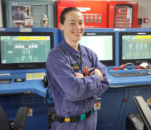Able Marine Technician (Electrical) Gemma Townshend hold some tools in her arms and is wearing  blue overalls. She stands in front of monitors in a control room.