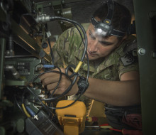 New Zealand Army's SGT Teaora Wright, an Electronics Technician working on electronics in a vehicle. He is wearing camouflage uniform and a head torch.