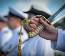 A sailor holds the handle of a ceremonial sword which has a gold handle, other sailors are blurred in the background.