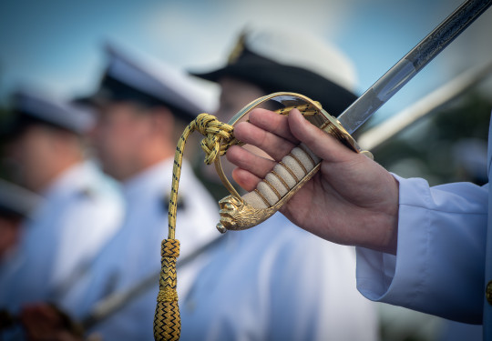 A sailor holds the handle of a ceremonial sword which has a gold handle, other sailors are blurred in the background.