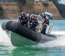 A RHIB travels towards the camera as it turns left with participants from School to Seas smiling on board. Those on the RHIB are wearing overalls, life jackets and helmets with visors. The water is very clear and cliffs are visible in the background.