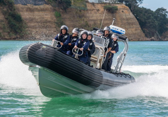 A RHIB travels towards the camera as it turns left with participants from School to Seas smiling on board. Those on the RHIB are wearing overalls, life jackets and helmets with visors. The water is very clear and cliffs are visible in the background.