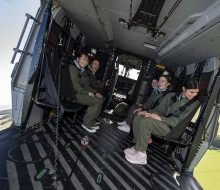 Five female high school students with wide smiles siting inside an NH90 helicopter during flight. Both the left and right side doors are open and the students are looking out and can see the green pastures below them. 