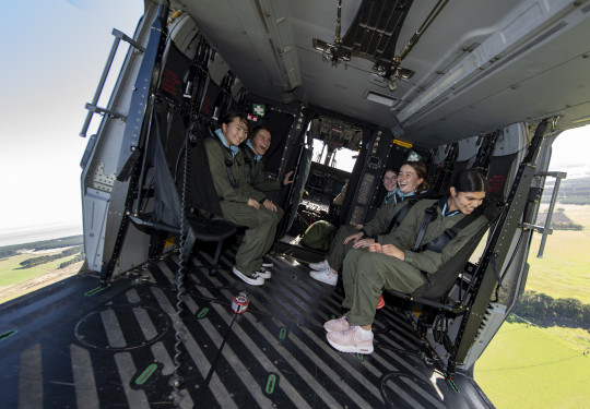 Five female high school students with wide smiles siting inside an NH90 helicopter during flight. Both the left and right side doors are open and the students are looking out and can see the green pastures below them. 