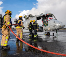 Four firefighters two in grey suits holding a red hose, and two in black suits, with yellow helmets tending to the shell of a Seasprite helicopter used in the exercise to simulate an aircraft crash.