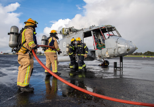 Four firefighters two in grey suits holding a red hose, and two in black suits, with yellow helmets tending to the shell of a Seasprite helicopter used in the exercise to simulate an aircraft crash.