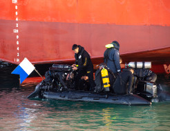 Divers, on a small boat prepare to dive in front of the the hull of a large ship.