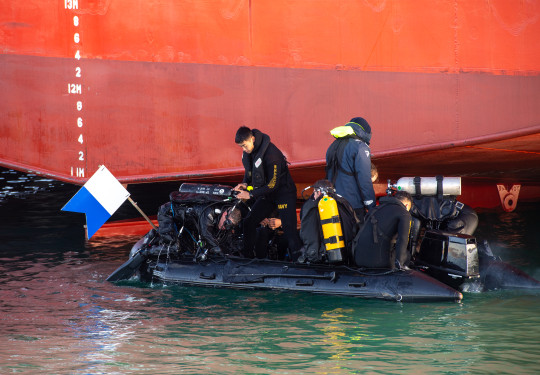 Divers, on a small boat prepare to dive in front of the the hull of a large ship.