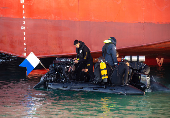 Divers, on a small boat prepare to dive in front of the the hull of a large ship.