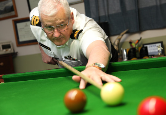 A low angle shot of a man lining up his cue against a white ball on a snooker table. The man is wearing a Navy uniform including a white shirt, black and gold rank slides, a name badge and a watch.
