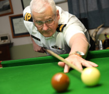 A low angle shot of a man lining up his cue against a white ball on a snooker table. The man is wearing a Navy uniform including a white shirt, black and gold rank slides, a name badge and a watch.
