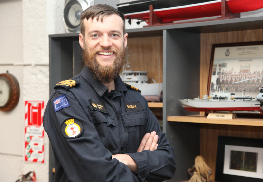 Lieutenant Commander Jonathan Otto wears a RNZN uniform and smiles at the camera. Behind him are models and pictures of ships in a wooden cabinet.