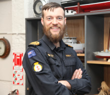 Lieutenant Commander Jonathan Otto wears a RNZN uniform and smiles at the camera. Behind him are models and pictures of ships in a wooden cabinet.