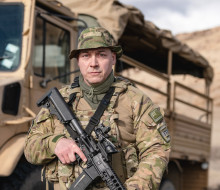 A soldier stands in uniform facing the camera holding a weapon with a New Zealand Army truck in the background