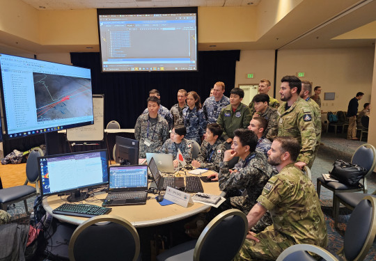 A number of uniformed military personnel stand in a room with lights on looking at various computer screens. 