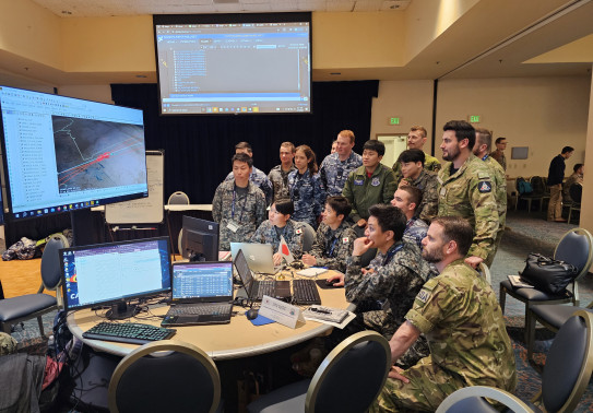 A number of uniformed military personnel stand in a room with lights on looking at various computer screens. 