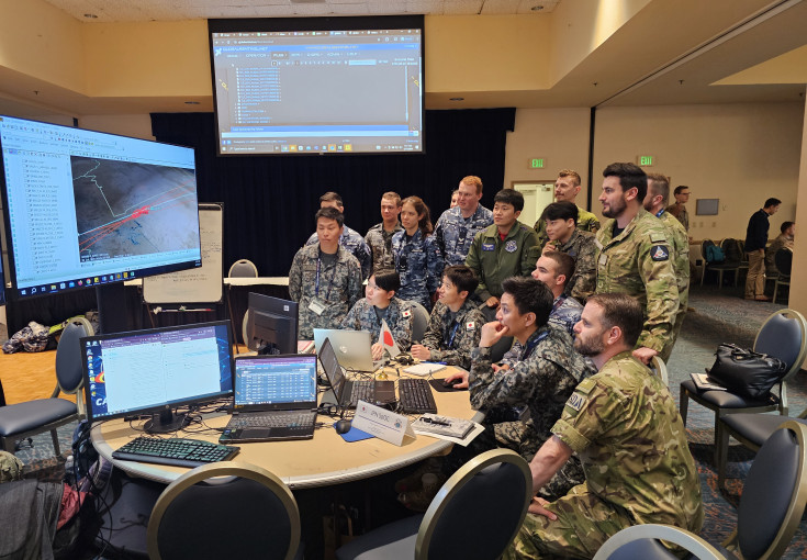 A number of uniformed military personnel stand in a room with lights on looking at various computer screens. 
