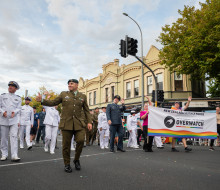 Sailors, Aviators and Soldiers walk through the streets of Auckland with a NZDF Overwatch Banner. They are smiling, waving and sharing a thumbs up with the crowd.