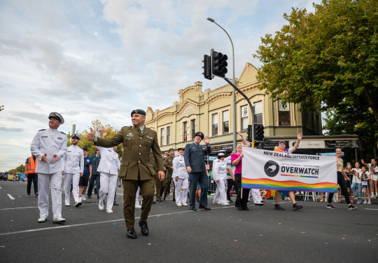 Sailors, Aviators and Soldiers walk through the streets of Auckland with a NZDF Overwatch Banner. They are smiling, waving and sharing a thumbs up with the crowd.