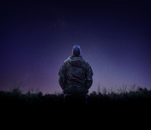 An aviator from the Royal New Zealand Air Force looks to the stars while wearing a camouflage jacket and a cap that has the letters 'RNZAF' on the back. 