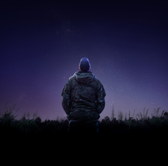 An aviator from the Royal New Zealand Air Force looks to the stars while wearing a camouflage jacket and a cap that has the letters 'RNZAF' on the back. 
