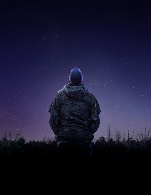 An aviator from the Royal New Zealand Air Force looks to the stars while wearing a camouflage jacket and a cap that has the letters 'RNZAF' on the back. 