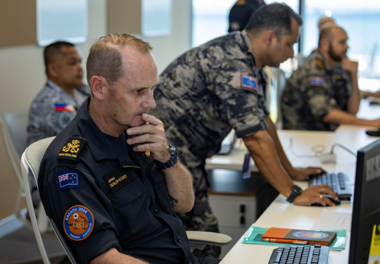 A sailor sits at a computer in a suit with other personnel working in the background.