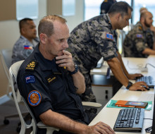 A sailor sits at a computer in a suit with other personnel working in the background.