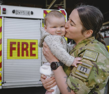 A soldier in uniform with eyes closed kisses baby who's looking at the camera. There's a fire truck in the background which says 'fire' on it.