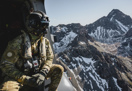 A helicopter loadmaster in full Air Force uniform including a flight helmet looks out the side of the aircraft to snow-capped mountains.
